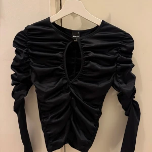 Svart långärmad topp från Gina Tricot - Snygg svart långärmad topp från Gina Tricot med rynkade detaljer och en stilren öppning framtill. Perfekt för en kväll ute eller en stilfull vardag.