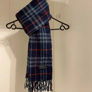 Burberry - Snygg rutig halsduk från Burberry i mörk blått med fransar. Den är i mycket fint skick, Perfekt för att hålla sig varm och stilren under kyliga dagar. Hör av er vid funderingar