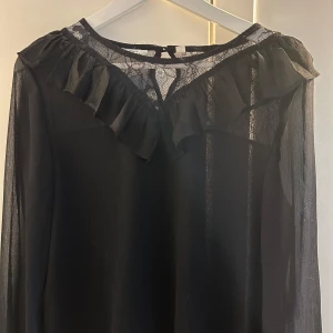 Svart blus med volangdetaljer - Elegant svart blus med volangdetaljer runt halsen. Blusen har långa ärmar och en transparent look som ger en sofistikerad stil. Perfekt för en chic och stilren outfit. Stl 164 men passar bra på både XS och S