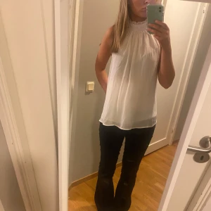 Vit ärmlös blus - Säljer en stilren vit blus med hög krage och ärmlös design. Perfekt för både vardag och festliga tillfällen. Blusen har en luftig passform och är lätt att matcha med olika outfits. (Använd en gång)