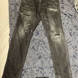 Dsquared2 jeans - Snygga svarta jeans från Dsquared2 med slitna detaljer och en cool, tvättad look. De har en klassisk femficksdesign och knappgylf. Perfekta för en trendig och avslappnad stil. Den är äkta och köpt från care of Carl några månader sen för 5000kr. Skriv om du har fler funderingar!