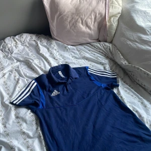 Blå pikétröja från Adidas - Snygg blå pikétröja från Adidas med klassiska vita ränder på ärmarna och logga på bröstet. Tröjan har korta ärmar och en bekväm passform. Aldrig använd, sällsynt modell! Skriv vid funderingar 💫✨