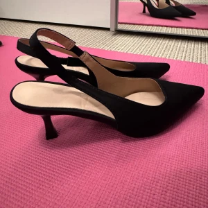 Svarta pumps med spetsig tå - Eleganta svarta pumps med spetsig tå och smal klack. Perfekta för en stilren look. Skorna har en öppen häl med rem för extra komfort.