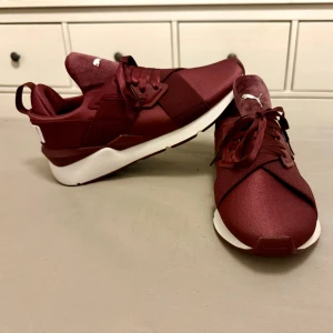 Vinröda sneakers från Puma - Snygga vinröda sneakers från Puma med vit sula och snörning. Skorna har en modern design med Pumas logga på  hälen. Perfekta för en sportig look.