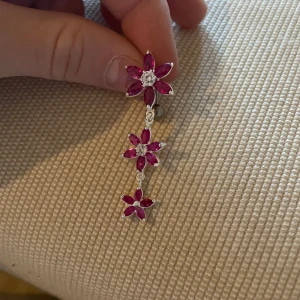 Blommig Navelpersing med rosa stenar - Snygg Navelpersing i silver med tre hängande blommor dekorerade med gnistrande rosa stenar. Perfekta för att ge en färgklick till din outfit. Blommorna är sammanlänkade och ger en elegant look.