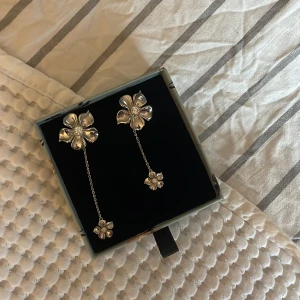 Silverfärgade blommiga örhängen - Eleganta silverfärgade örhängen med blommotiv. De har en lång kedja som förbinder två blommor, vilket ger en stilren och modern look. Perfekta för att lyfta vilken outfit som helst.