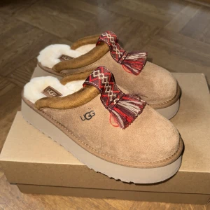 Helt nya Bruna/Beiga UGG Tazzle  - Säljer ett par supermysiga och sköna bruna tofflor från UGG i mocka med liten tofs. Insidan är fodrad med mjuk mysig päls. Väldigt sköna! 