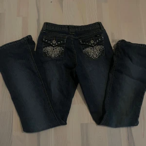 Mörkblå jeans med broderade bakfickor - Snygga mörkblå jeans med unika broderade detaljer och pärlor på bakfickorna. Perfekta för att ge din outfit en extra touch. Klassisk femficksdesign med knapp och dragkedja framtill.