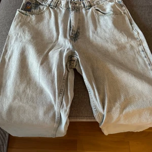 Ljusa jeans Big Boy Byxor - Säljer ett par ljusa baggy jeans från 99 Denim med en cool broderad detalj på fickan. Byxorna har en loose passform och är perfekta för en avslappnad stil. De har en klassisk femficksdesign och knappgylf. 