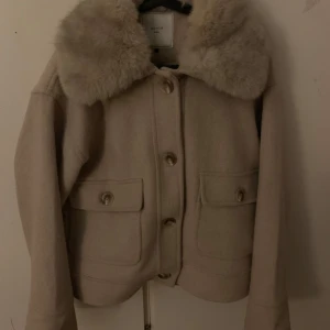 Beige kappa med pälsdetaljer från Neo Noir - Snygg beige kappa från Neo Noir med stora pälsdetaljer på kragen. Jackan har knappar framtill och två fickor med lock. Perfekt för kyligare dagar när du vill vara både varm och stilren.