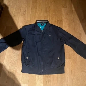 Mörkblå bomberjacka från GANT - Snygg mörkblå bomberjacka från GANT med dragkedja och ribbade muddar. Jackan har två fickor med knappar och ett diskret GANT-märke på bröstet. Perfekt för en stilren look. skriv privat för bild på 