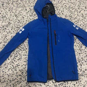 Blå vindjacka från Sail Racing - Snygg blå vindjacka från Sail Racing med huva och dragkedja. Jackan har en bröstficka med dragkedja och logotypdetaljer på ärmen. Perfekt för blåsiga dagar.