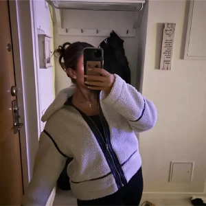 teddyjacka - Mysig vit teddyjacka med svarta detaljer och dragkedja framtill. Perfekt för kyliga dagar med sin fluffiga och bekväma design. Jackan har långa ärmar och en avslappnad passform.