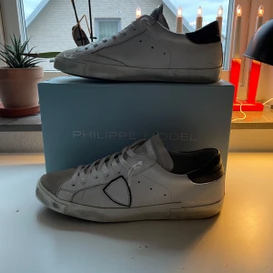 Philippe Model PM/78EDT skor - Snygga vita sneakers från Philippe Model med svarta detaljer. Skorna har en klassisk design med snörning och en diskret logotyp på sidan. Perfekta för en stilren look.