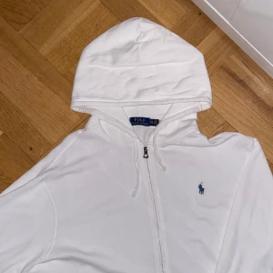 Vit hoodie från Polo Ralph Lauren - Säljer en stilren vit hoodie från Polo Ralph Lauren med dragkedja och den klassiska loggan broderad i blått på bröstet. Perfekt för en avslappnad look.