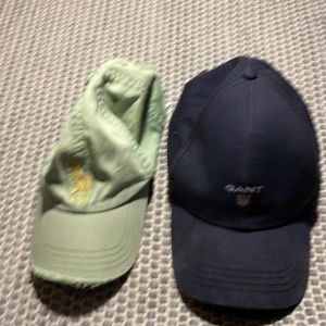 Två kepsar från Polo Ralph Lauren och GANT - Säljer två stilrena kepsar. En grön keps från Polo Ralph Lauren med broderad logga och en mörkblå keps från GANT med broderad logga framtill. Perfekta för en avslappnad stil.