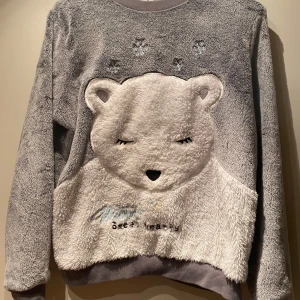 Grå fluffig tröja med isbjörn motiv st XS - Mysig grå tröja med ett stort isbjörnansikte i fluffigt material på framsidan. Tröjan har långa ärmar och ribbade muddar. Perfekt för kalla dagar när du vill känna dig extra bekväm och söt. Snöflingor i ljusblå och text som det står Winter sweet hearts. Super mjuk och gosig. St XS från Cubus. 