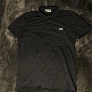 Svart pikétröja från Lacoste - Säljer en klassisk svart pikétröja från Lacoste med korta ärmar och den ikoniska krokodilloggan på bröstet. Tröjan har en stilren krage och knappar framtill. Perfekt för en avslappnad men stilfull look.