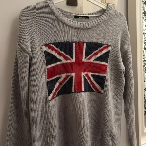 Grå stickad tröja med Union Jack från Gina Tricot - Säljer en snygg grå stickad tröja från Gina Tricot med ett stort Union Jack-motiv på framsidan. Tröjan har långa ärmar och ribbade kanter vid ärmslut och nederkant. Perfekt för en cool och avslappnad stil. 🖤
