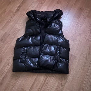 Svart dunväst med huva - Snygg svart dunväst med glansig yta och praktisk huva. Västen har en dragkedja framtill och två stora fickor. Perfekt för kyliga dagar när du vill hålla stilen.