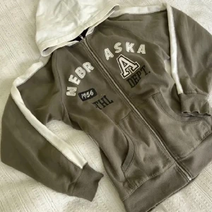 Grön och vit hoodie med Nebraska-tryck - Lånade bilder!! Säljer en  olivgrön zip up hoodie med vit huva och detaljer!  Köptes här på plick men kommer inte till användning , inga defekter 