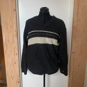 Mörkgrå tröja med v-ringning - Säljer en mörkgrå tröja med v-ringning och ett beige randigt mönster över bröstet. Tröjan har långa ärmar och en avslappnad passform, perfekt för kyligare dagar. 