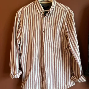 Randig skjorta från Jack & Jones - Snygg randig skjorta från Jack & Jones i vitt och vinrött. Skjortan har långa ärmar och knappar framtill samt en bröstficka. Storlek L 