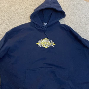Mörkblå hoodie från Sweet SKTBS - Säljer en mörkblå hoodie från Sweet SKTBS med en cool broderad logga i gult och vitt på framsidan. Den har en klassisk känguruficka och justerbar huva. Perfekt för en avslappnad stil!