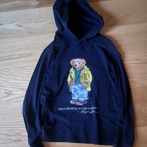 Ralph Lauren hoodie - Säljer en marinblå hoodie från Ralph Lauren med det ikoniska Polo Bear-motivet på framsidan. Björnen bär en jacka och blå jeans, vilket ger en cool och avslappnad look. Perfekt för en casual stil! Den är storlek Y-L, men passar som XS