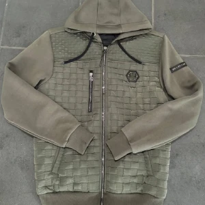 Grön jacka med huva från Philipp Plein - Snygg grön jacka med huva från Philipp Plein. Jackan har ett quiltat mönster på framsidan och en dragkedja i mitten. Den har också en cool logotypdetalj på bröstet och en ärm. Perfekt för en stilren look! Skick 9/10