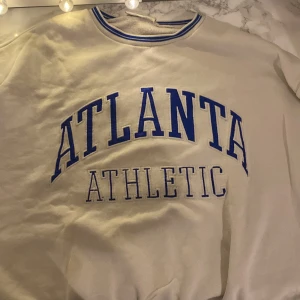 Vit sweatshirt från Chiquelle - Säljer en vit sweatshirt från Chiquelle med blå text 'ATLANTA ATHLETIC' på framsidan och en stor blå siffra '8' på baksidan. Tröjan har en rund halsringning med blå ränder. Mudden är förstörd på ena ärmen.