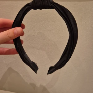 Svart diadem med knutdetalj - Snyggt svart diadem med en elegant knutdetalj på toppen. Perfekt för att ge en stilren touch till din look. Materialet har en lätt glansig finish som ger en extra dimension.