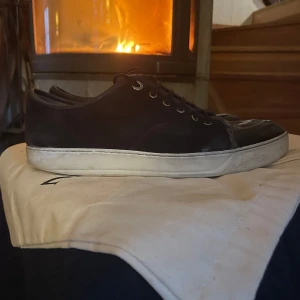 Navy blue captoe sneakers från Lanvin - Snygga svarta sneakers från Lanvin med snörning och en stilren design. Skorna har en kombination av mocka och skinn, vilket ger dem en unik look. Skicket är 7/10. Defekter är på toeboxen och insidan av skon, (går nog att reperara) Storleken är UK 9 vilket motsvarar 43-44 men de sitter stort i storleken. Tillbehör är 2 dustbags, ingen orginal box tyvärr.