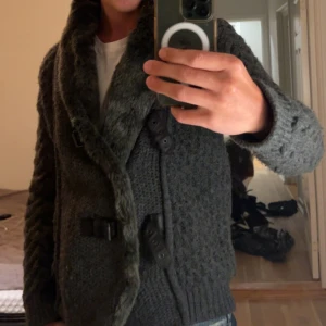 Grå stickad kofta med pälsdetaljer - Säljer en grå stickad kofta med pälsdetaljer på kragen. Koftan har ett unikt mönster och stängs med spännen framtill. Perfekt för kyliga dagar.