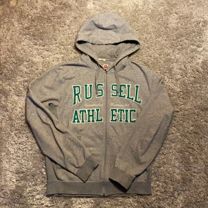 Grå zip hoodie - Säljer en grå zip hoodie från Russell Athletic med dragkedja och gröna bokstäver på framsidan. Tröjan har en justerbar huva och långa ärmar med ribbade muddar. Perfekt för en avslappnad stil.