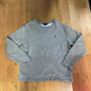 Grå sweatshirt från Polo Ralph Lauren - Säljer en stilren grå sweatshirt från Polo Ralph Lauren med det klassiska broderade logotypen på bröstet. Tröjan har långa ärmar och en bekväm passform, perfekt för en avslappnad look. Passar bra till både jeans och chinos. 