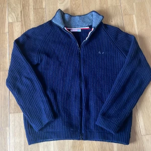 Mörkblå stickad cardigan från SUN68 - Säljer en mörkblå stickad cardigan från SUN68 med dragkedja och grå ribbad krage. Den är i storlek L men passar mer som M. Den är perfekt för kyliga dagar! Tveka inte att höra av dig vid frågor och funderingar!😁💯