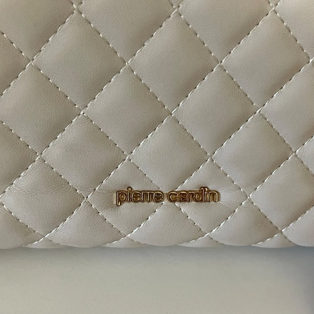 Snygg vit handväska från Pierre Cardin med quiltat mönster och guldfärgade kedjedetaljer. Väskan har en elegant spänne framtill och justerbar axelrem. Perfekt för att ge din outfit en lyxig touch.. Laukut & Käsilaukut.