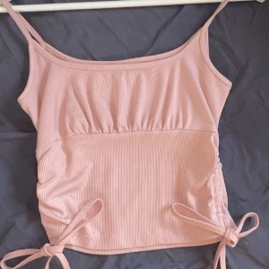 Rosa ribbad linnetopp - Säljer en söt rosa ribbad linnetopp med smala axelband och knytning i sidorna. Perfekt för varma dagar och kan enkelt matchas med olika outfits. Toppen har en enkel och stilren design.