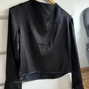 Svart blus med vita sömmar - Snygg svart blus med kontrasterande vita sömmar. Jackan har långa ärmar och en stilren design. Perfekt för en minimalistisk look.