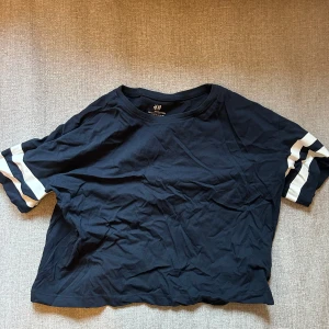 Mörkblå croppad t-shirt från H&M - Säljer en mörkblå t-shirt från H&M i  bomull. T-shirten har korta ärmar med vita ränder och en avslappnad passform. Perfekt för en casual look!