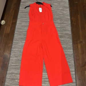 Röd ärmlös jumpsuit BIK BOK - Snygg röd ärmlös jumpsuit med djup v-ringning och markerad midja. Perfekt för en stilren look. Den har vida ben och en dragkedja i ryggen för enkel påklädning.