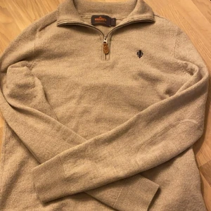 Beige merinoulltröja från Morris - Säljer en stilren beige tröja från Morris i 100% merinoull. Tröjan har en dragkedja vid halsen och en broderad detalj på bröstet. Perfekt för kyligare dagar med sin mjuka och varma ullkvalitet. Det är storlek S men har krympt i tvätten så skulle uppskatta xs/165