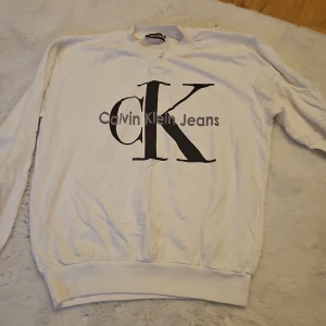 Vit tröja från Calvin Klein - Vit tröja från Calvin Klein med stort 'CK' tryck på framsidan. Tröjan har långa ärmar och en klassisk rund halsringning. Perfekt för en stilren och avslappnad look.