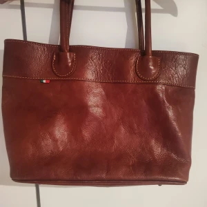 Brun äkta skinnväska från Vera Pelle - Säljer en snygg konjak skinnväska från Vera Pelle. Väskan har en praktisk dragkedja på baksidan och rymligt innerfack med ljus textilfoder. Perfekt för att bära med sig det viktigaste med stil. Made in Italy för en touch av elegans. Helt oanvänd. 