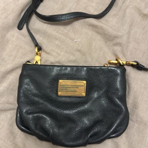 Svart axelväska från Marc by Marc Jacobs - Snygg svart axelväska i skinn från Marc by Marc Jacobs med guldfärgade detaljer. Väskan har en justerbar axelrem och en dragkedja upptill. Insidan är fodrad med ett mönster av märkets logotyp. Perfekt för att bära dina nödvändigheter med stil.