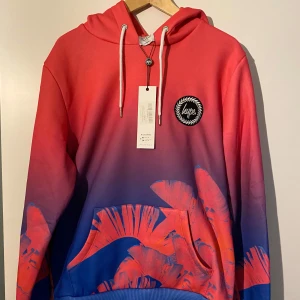Färgglad hoodie från Hype - Säljer en färgglad hoodie från Hype med en cool ombre-effekt i rosa och blått. Tröjan har en stor ficka framtill och justerbar huva med vita snören. Perfekt för att sticka ut i mängden! Oanvänd, endast provad.