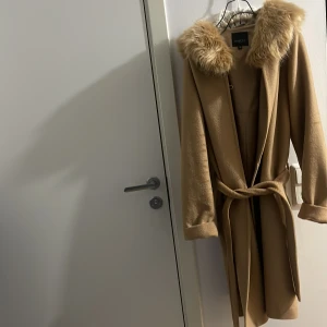Beige kappa från Hollies - Säljer en elegant beige kappa från Hollies med en lyxig fuskpälsdetalj runt kragen. Kappan har ett knytbälte i midjan och långa ärmar, perfekt för kyliga dagar. En stilren och tidlös design som passar till många tillfällen. Har köpt den för 5800kr för 6 månader sedan och har använts endast en gång