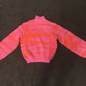 Rosa och orange tröja från Crās - Säljer en färgglad och fluffig tröja från Crās i rosa och orange. Tröjan har en hög krage och långa ärmar, perfekt för att lysa upp garderoben. Den är randig och har en mysig känsla. Passar bra för kyligare dagar.