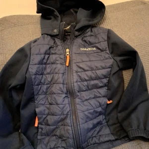 Blå jacka från Didriksons - Säljer en blå jacka från Didriksons med quiltad framsida och dragkedja. Jackan har en huva och långa ärmar, perfekt för kyligare dagar. Den har även orange detaljer vid dragkedjan och fickorna.